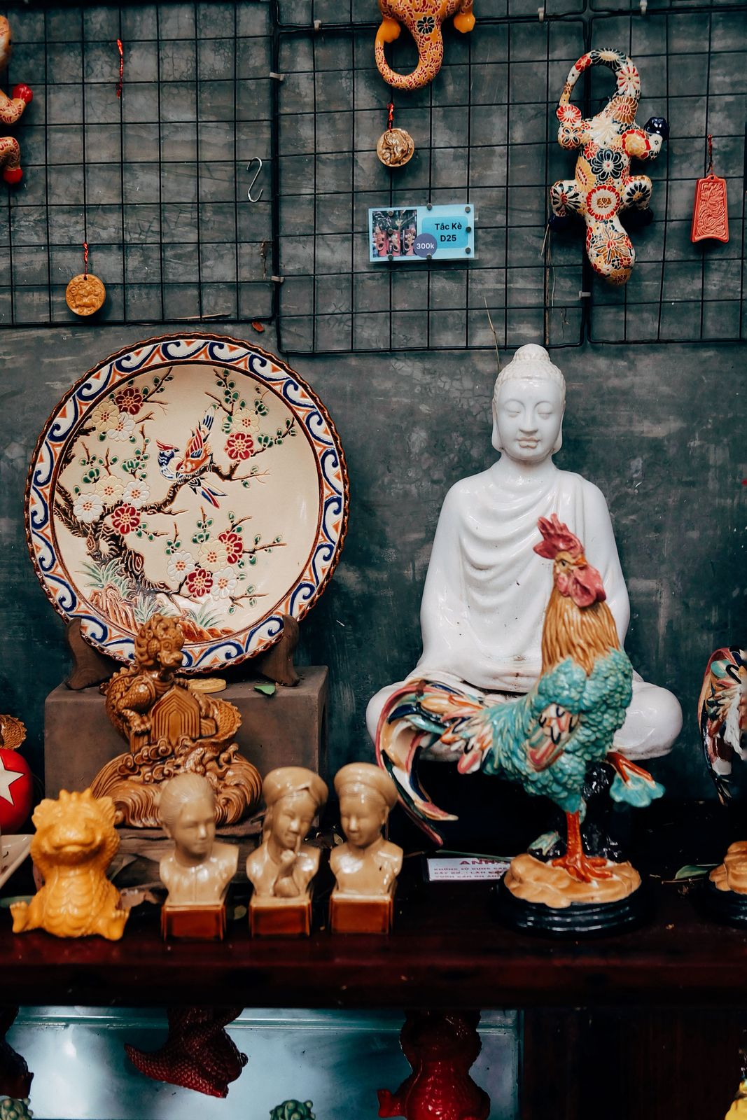 Glazed ceramic rooster in turquoise and amber standing before a white seated meditation statue, with a painted blossom plate and small busts