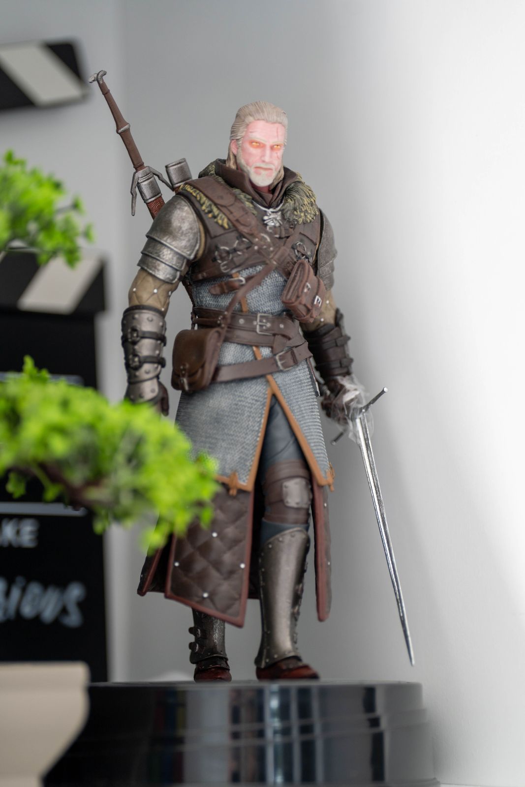Painted statue of a silver-haired swordsman in chainmail and leather armor holding a long sword, beside a small bonsai